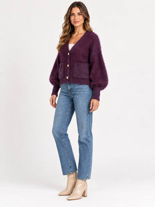 A woman modeling a fuzzy, shaggy purple cardigan with front pockets and gold buttons, paired with blue jeans and beige ankle boots, against a plain white studio background.