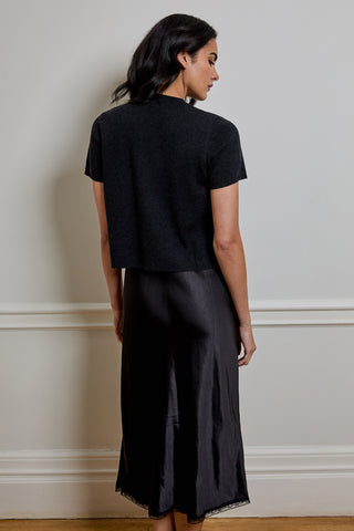 A woman shown from the back wearing a black short-sleeve knit top and a black satin midi skirt, standing against a light beige wall with white trim.