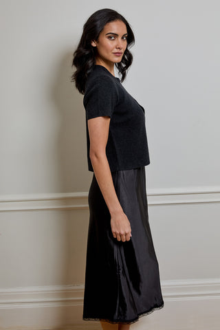 A woman wearing a black short-sleeve knit top and a black satin midi skirt, standing in profile against a light gray wall with white trim.