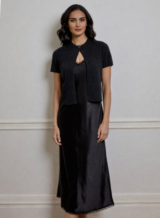 A woman modeling a black short-sleeve knit cardigan layered over a black satin midi dress, standing against a light gray wall with white trim.