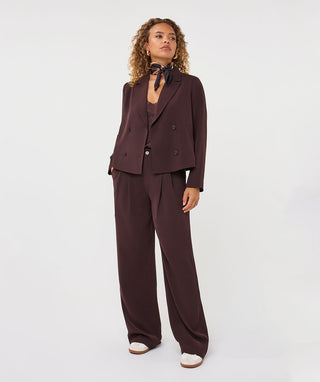 A model stands against a plain light gray studio background wearing a dark brown tailored suit set with a double-breasted blazer, matching vest, wide-leg trousers, a dark bow tie, and white shoes.
