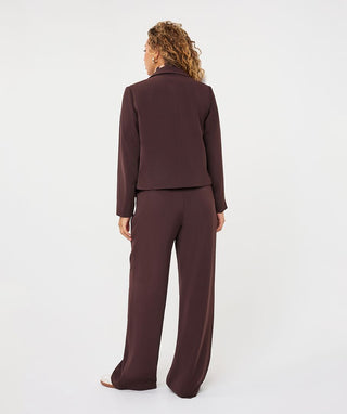 A woman viewed from the back wearing a matching dark plum-brown two-piece suit with a cropped blazer and wide-leg trousers, photographed against a clean white studio background.